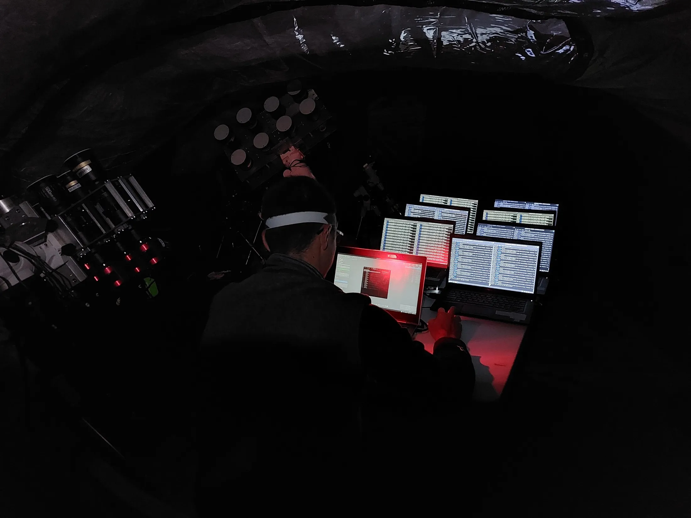 A researcher at field computers inside a dark tent, with instruments beside the screens