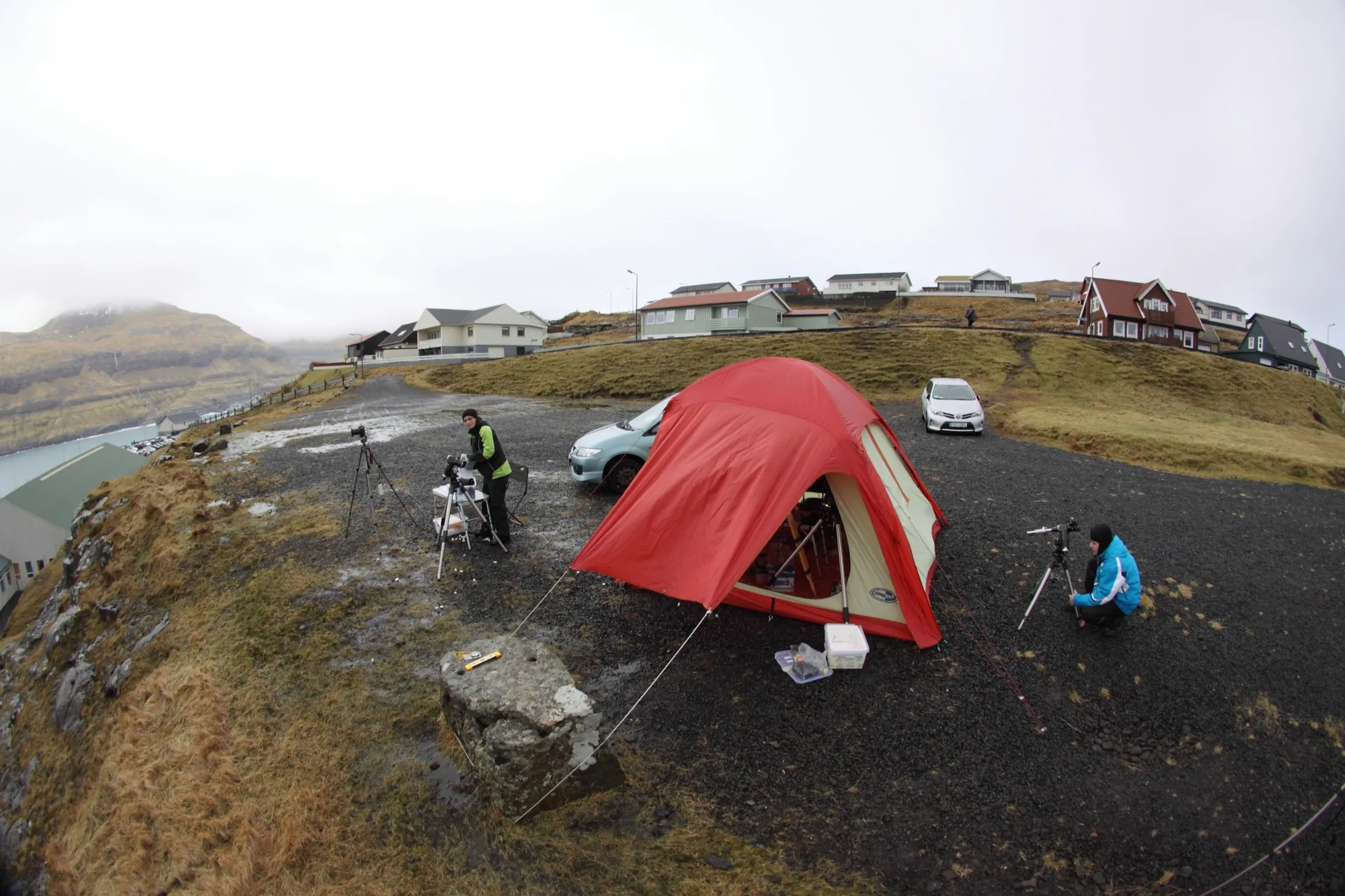 A red expedition tent and instruments on a coastal gravel site, with two people setting up tripods