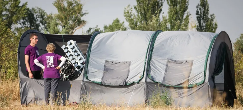Two team members in purple expedition shirts at the Burgos field camp, beside grey observation tents