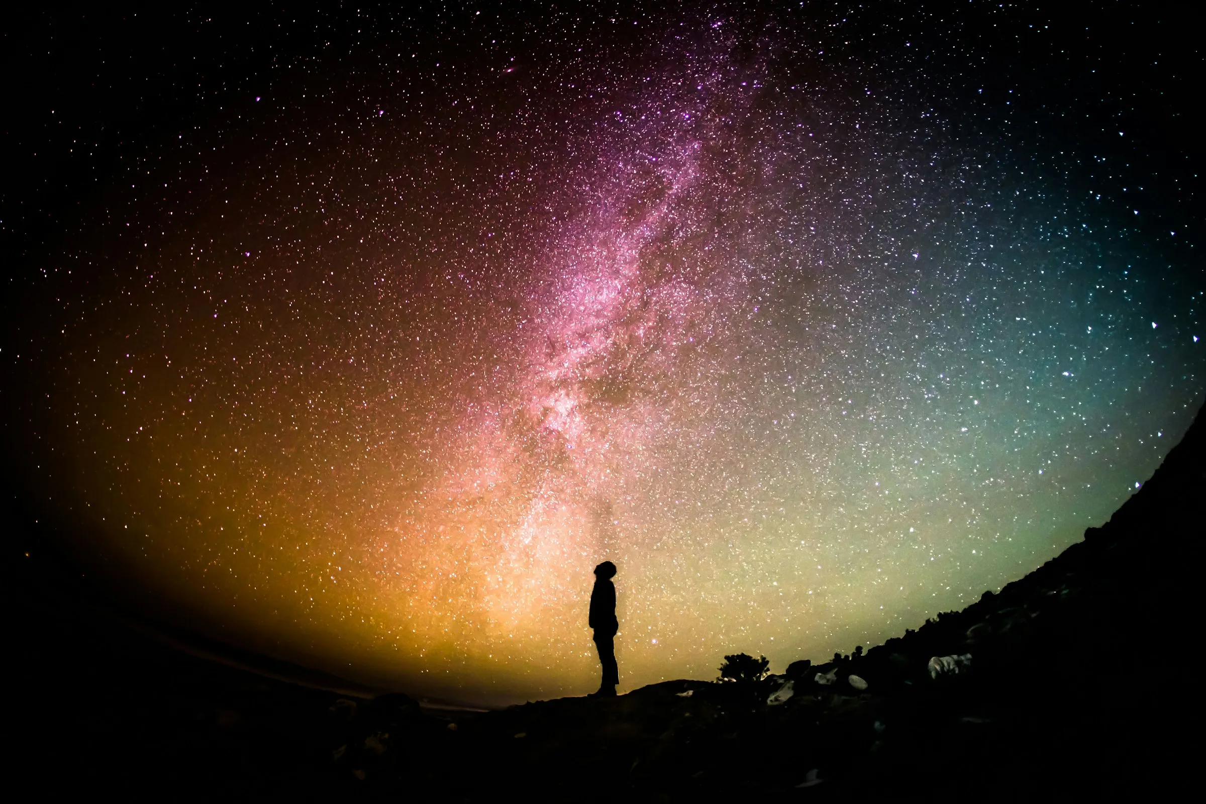 A lone figure stands on a dark ridge beneath the Milky Way, looking up at a densely starred night sky